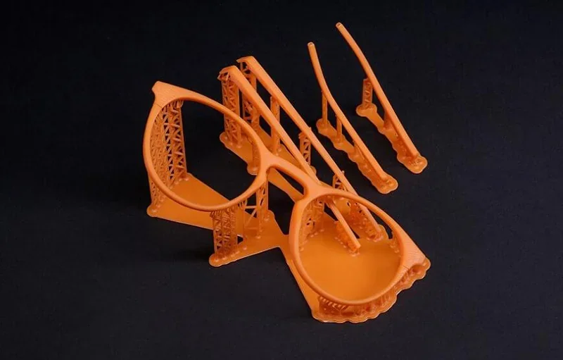 Orange 3D-printed eyeglass frame and parts with intricate supports, created by Prusa SL1S, displayed on a black background.