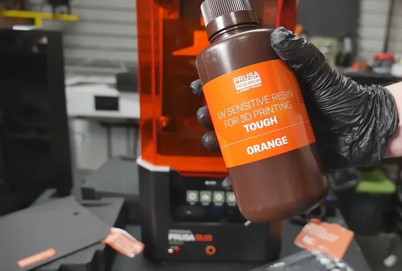 Prusa SL1S 3D printer with UV resin bottle labeled "Tough Orange" held by a gloved hand in a workshop setting.