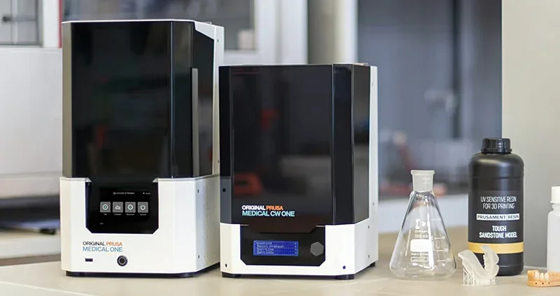 Prusa SL1S 3D printer with resin, flasks, and printed parts on a table in a lab setting.