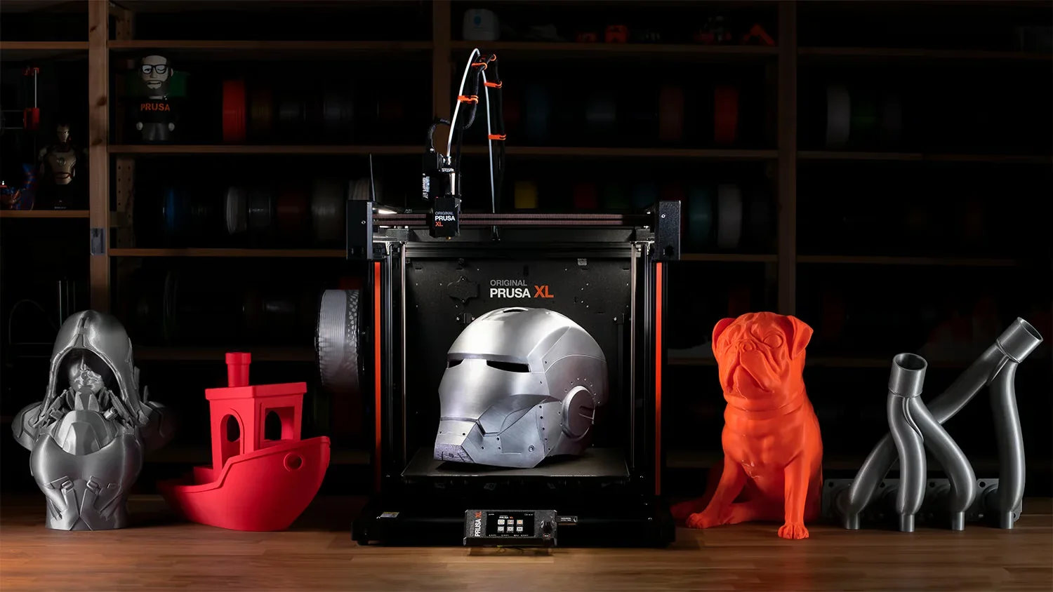 Prusa XL in a workshop with a printed helmet, red boat, orange dog, and silver parts showcasing its 3D printing capabilities.