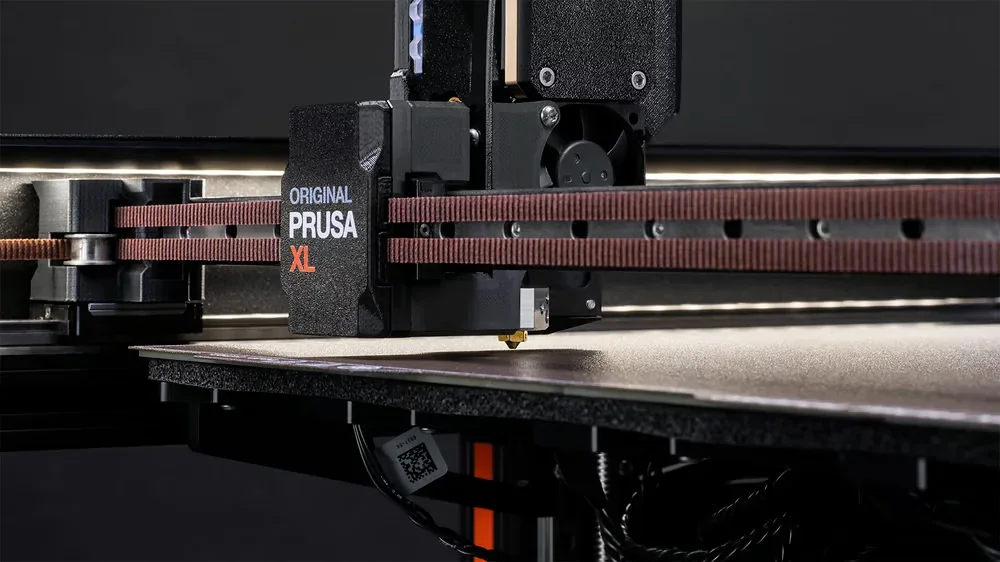 Prusa XL 3D printer detail, branded extruder head on a belt system, sleek design, partial view of bed surface, focused on printing components.