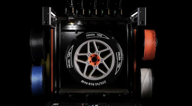 Prusa XL 3D printer with a printed wheel and tire labeled "Prusa Tire" surrounded by colorful filament spools in a dark setting.