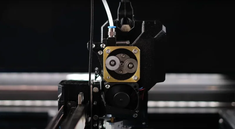 Prusa XL 3D printer head close-up with gears, nozzle, and tubing, showcasing precision engineering with a focus on high-quality part production.