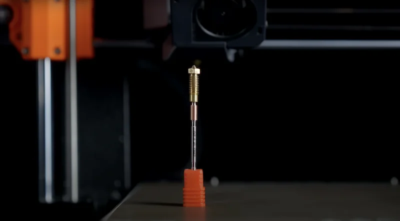 Prusa XL 3D printer detail showing nozzle assembly above a red 3D-printed base. Black background highlights precision engineering.