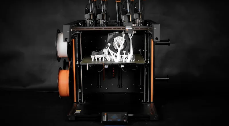 Prusa XL 3D printer with skeletal structure sample being printed, orange and black design, multiple extruders, visible filament spools.
