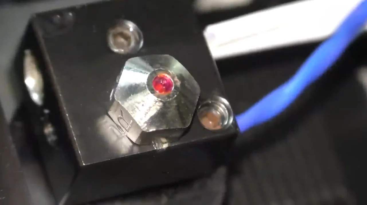 Qidi I-Fast 3D printer nozzle close-up, showing a shiny metallic part with a red center, blue wire attached, indicating precision engineering.