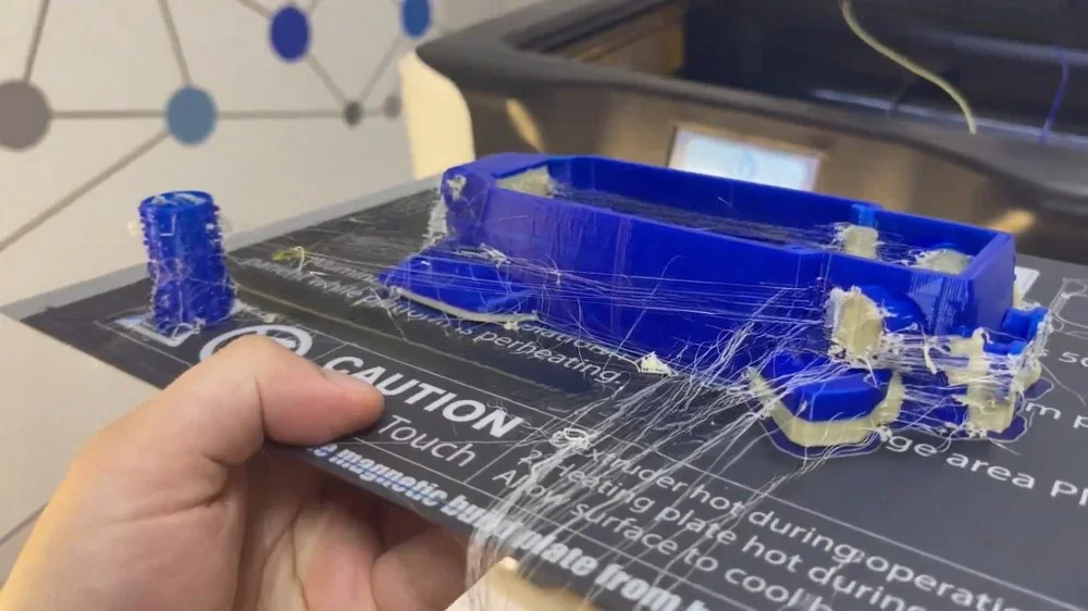 qidi i-fast 3D printer's blue prints with rough finish, stray strands, held above printer. Printed samples show potential quality issues.
