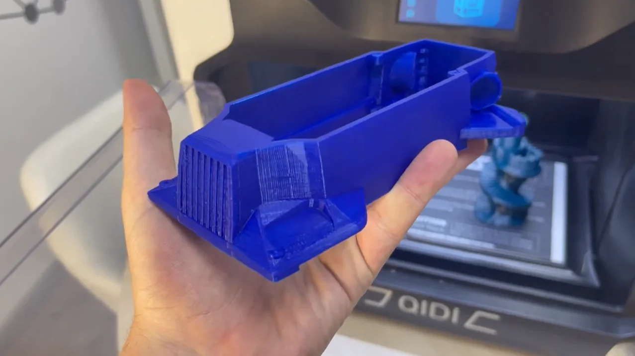 Blue 3D-printed model car part held in hand, created by Qidi I-Fast printer, with printer interior visible in background.