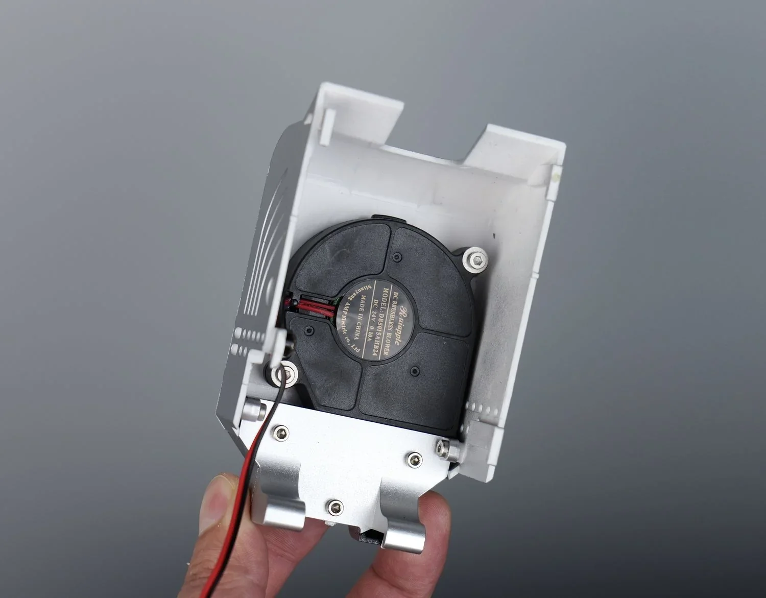 Qidi X Max 3D printer component with black fan, wires, and metal housing. Held in hand, showing design detail against gray background.