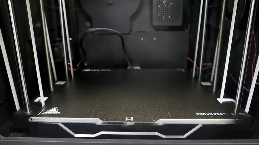 Qidi X Max 3D printer interior showing a spacious build platform with metal supports, black base, and visible wiring, ready for model creation.