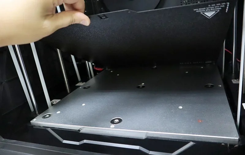 Qidi X Max 3D printer interior with a hand adjusting the build plate; sleek metallic finish and visible springs, no samples shown.