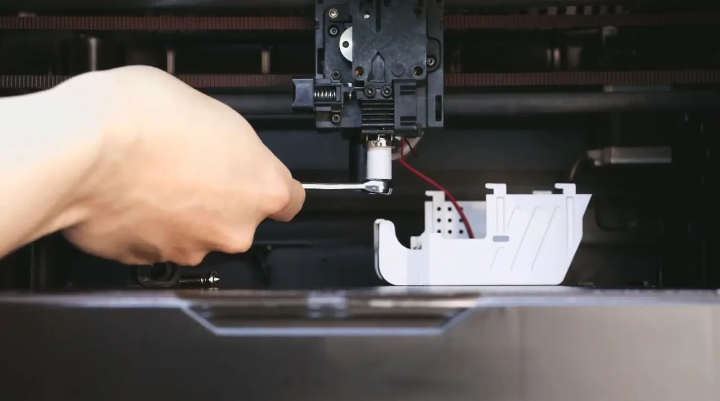 Qidi X Max 3D printer nozzle adjusted by a hand, with a partially printed white object visible, showcasing precise detail capabilities.