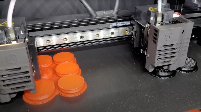 Raise3D E2 3D printer dual extruding circular orange parts on black surface.