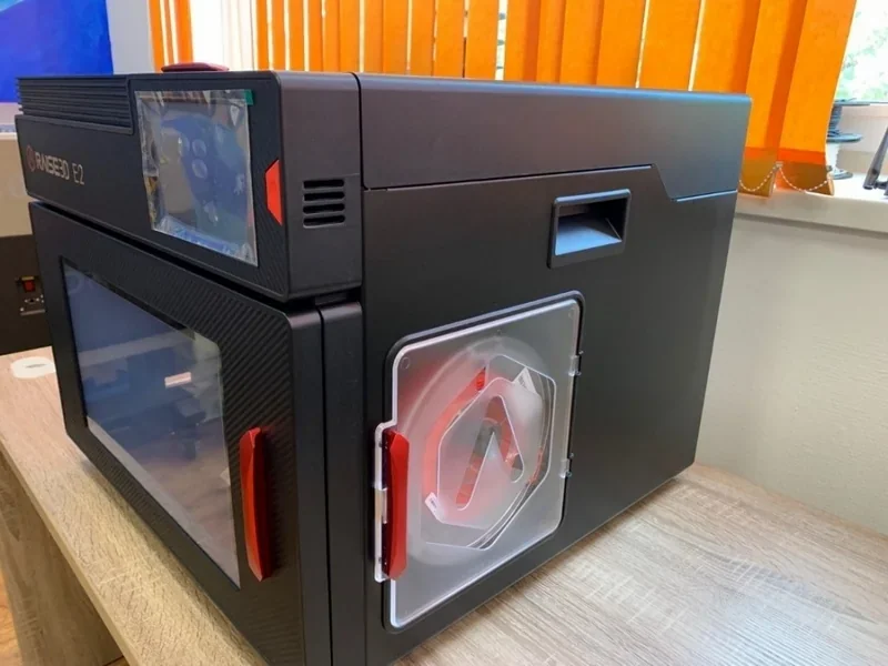 Raise3D E2 3D printer on a desk with black and red design, clear viewing window, and visible branding, positioned in a brightly lit room.