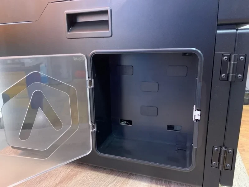 Raise3D E2 3D printer with an open side panel, revealing interior space, branding visible on transparent door, set on a wooden surface.