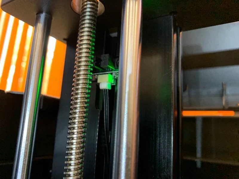 Raise3D E2 internal mechanism with rods, screw, and glowing green component, illuminated by orange light; part of a 3D printer setting.