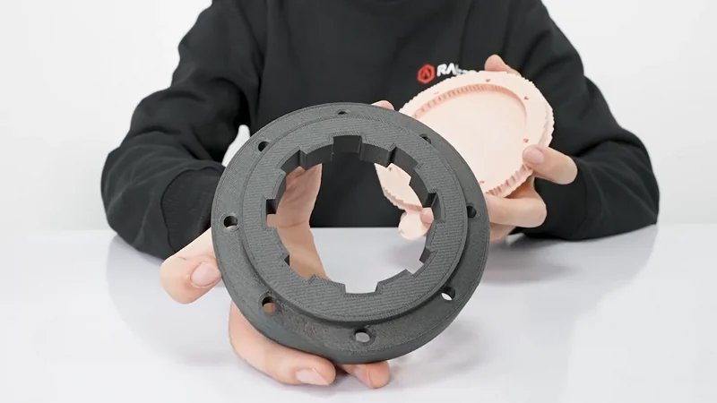 Person holds black and pink 3D printed parts from the Raise3D E2CF printer, wearing a black sweater with Raise3D branding in a white background setting.
