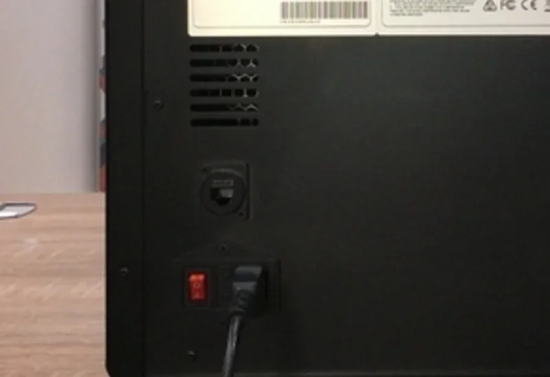 Back of a Raise3D E2CF printer showing vent, power switch, and connection port on a wooden table.