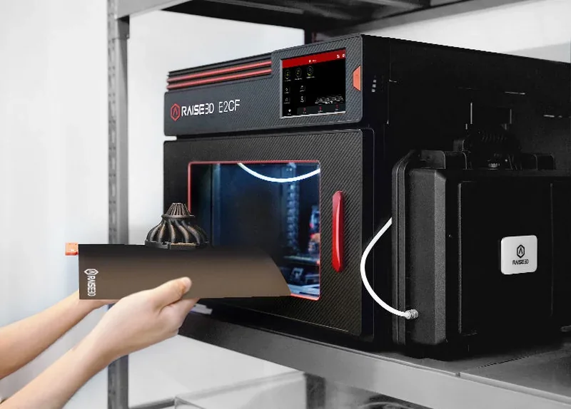 Raise3D E2CF 3D printer with carbon-fiber printed part, touchscreen interface, and branding visible on a shelf.