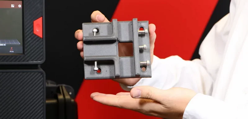 Raise3D E2CF 3D printer showing a printed gray mechanical part with bolts, held by a person in front of the printer with a red and black background.