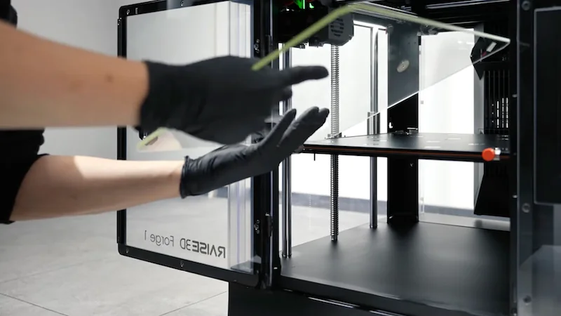 Raise3D MetalFuse with open chamber, person wearing gloves adjusting element, showcasing printer setup and interior for part creation.