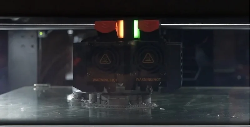 Raise3D MetalFuse 3D printer in action, dual extruders creating a part, illuminated by orange and green lights, displaying precision technology.