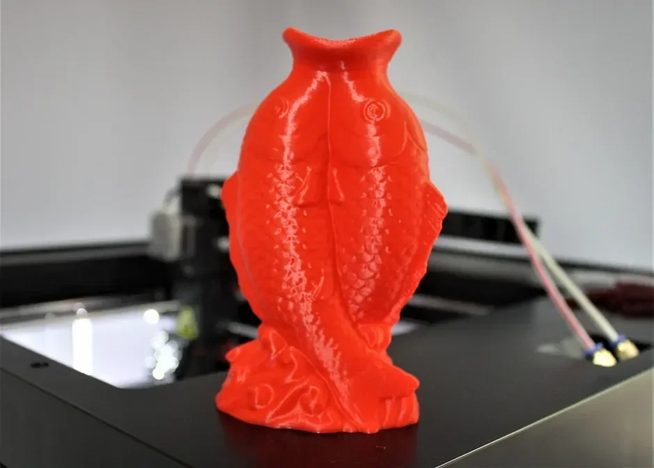 Red fish sculpture 3D printed by Raise3D Pro2 Plus, displayed on printer bed with background wiring and mechanical components visible.