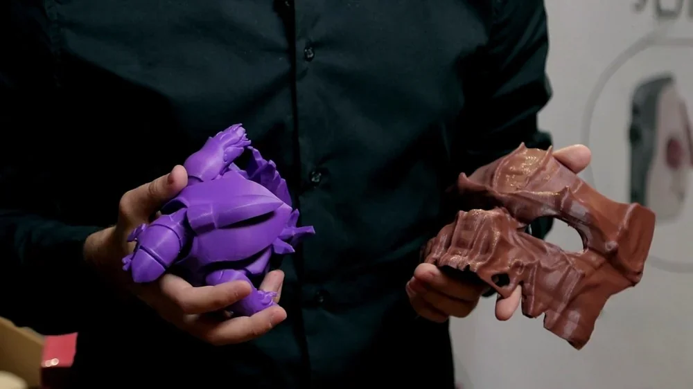 Hands holding 3D-printed models in purple and brown, crafted by Raise3D Pro2 Plus, showcasing its precision and capabilities in a workshop setting.