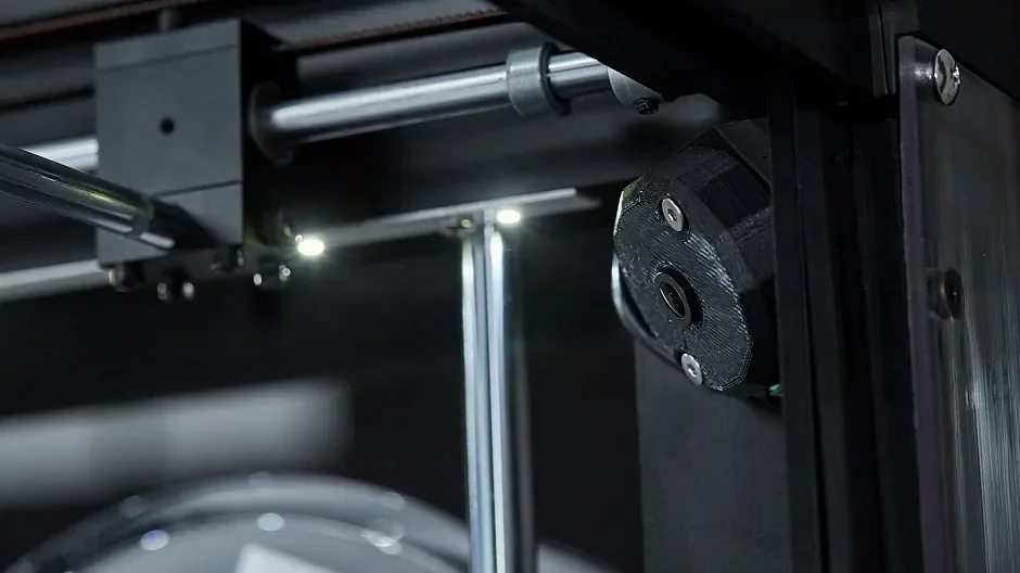Raise3D Pro2 Plus 3D printer close-up shows internal mechanics, including metal rods and a stepper motor, highlighting precision engineering.