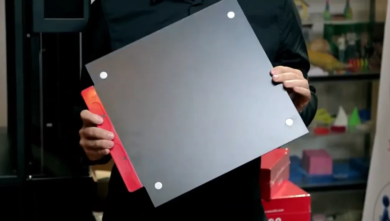 Raise3D Pro2 Plus print bed displayed in hands, surrounded by 3D printed models in a workshop setting.