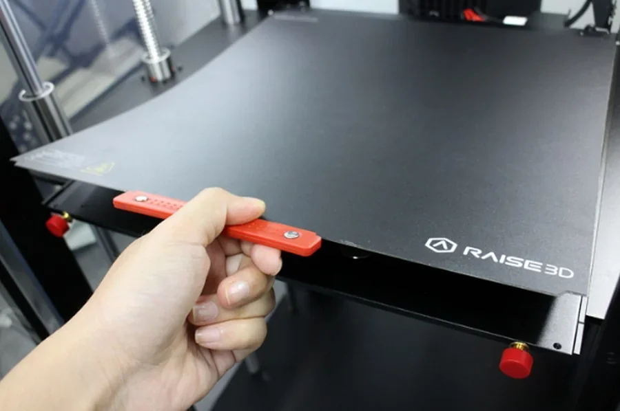 Raise3D Pro2 Plus 3D printer with a detachable build platform, visible black surface, hand holding red handle, showcasing brand logo.