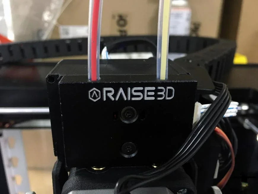 Raise3D Pro2 Plus 3D printer extruder with branding visible, wires and filament tubes connected, on an industrial shelf setting.