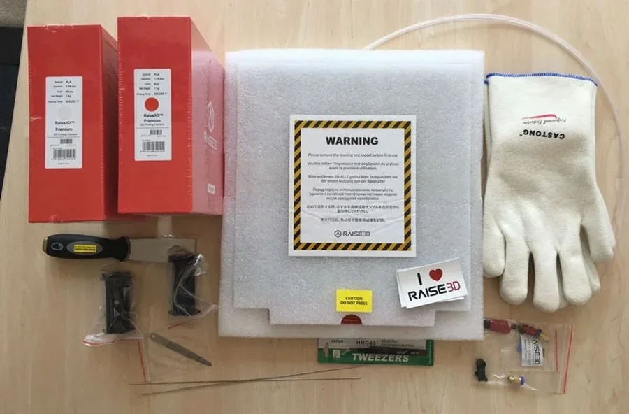 Raise3D Pro2 Plus accessories on a table, including gloves, spools, tweezers, scraper, warning foam, and branded boxes.