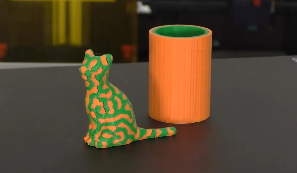3D printed objects: a colorful green and orange cat figurine and an orange cylinder, created by Raise3D Pro3, on a table surface.