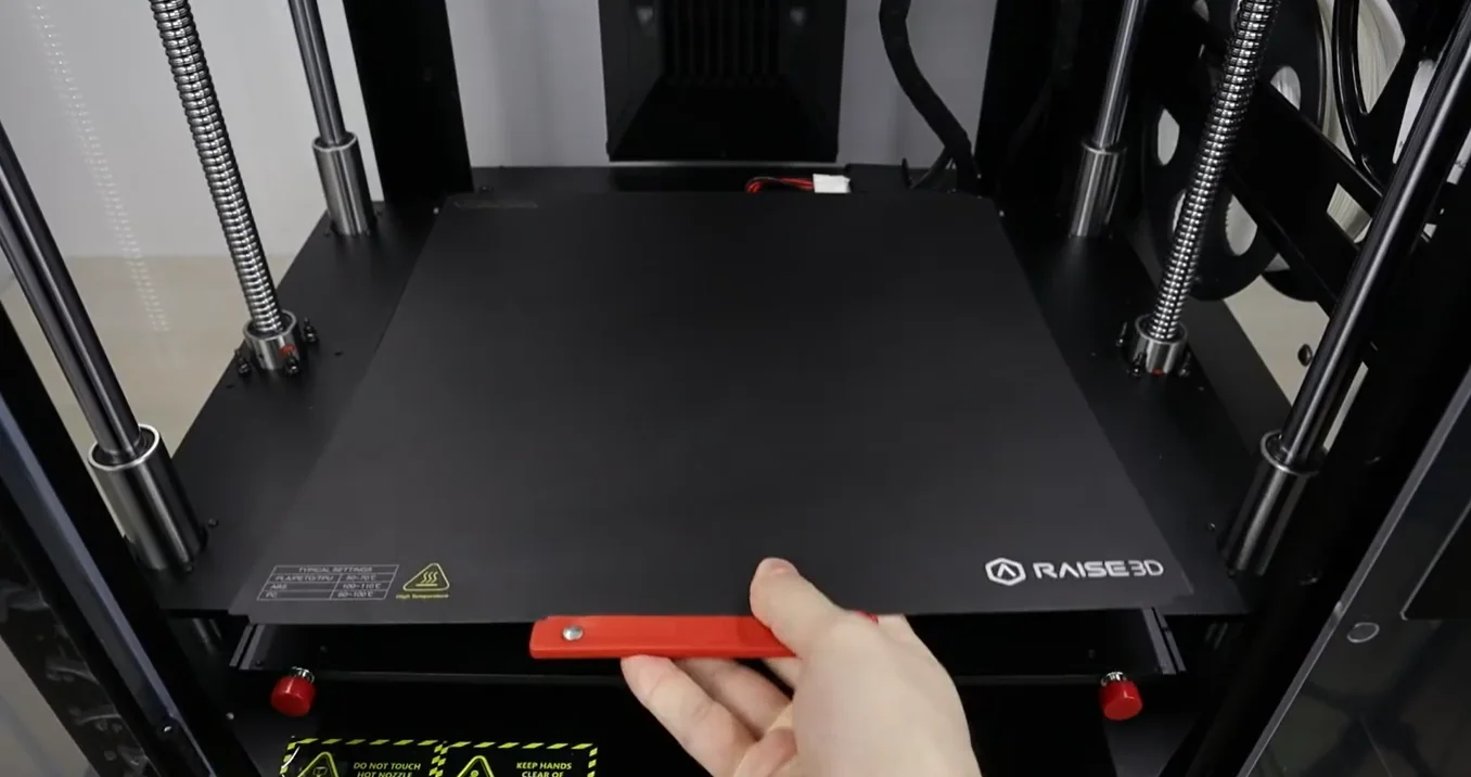 Hand adjusting the build plate in Raise3D Pro3 printer, featuring branding and robust mechanical components for precise 3D printing.