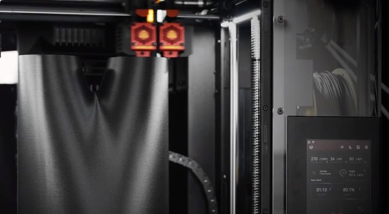 The image shows the interior of a Raise3D Pro3 HS 3D printer during operation. A large, black print object is being created inside the printer. The dual extruder system visible at the top features two nozzles, indicating its capability for multi-material or color printing. The printer's build chamber is well-lit and enclosed. On the right side, a digital display screen shows live printing statistics, including temperature, time, and completion percentage, enhancing user control and monitoring.