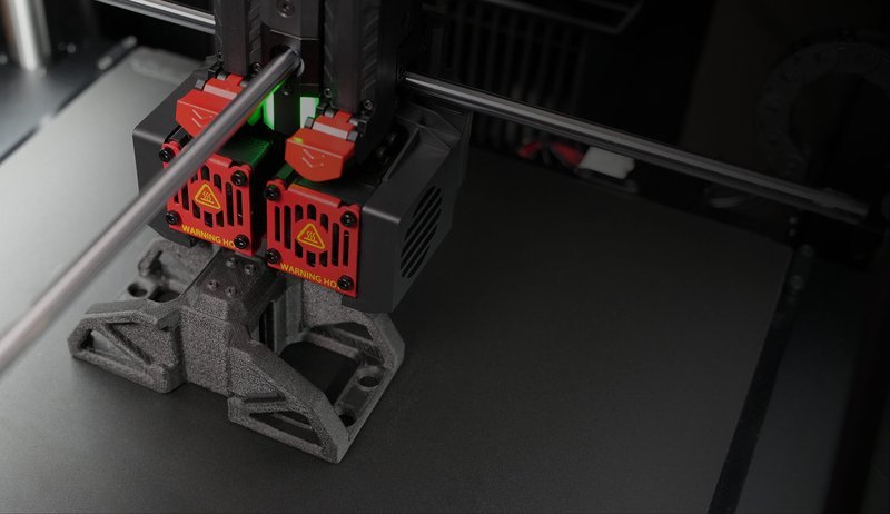 The image showcases the Raise3D Pro3 HS 3D printer in operation. The machine is actively printing a complex geometric part, with dual extrusion heads prominently visible. The print heads, marked with red and black protective housings and warning labels, are positioned over a textured build plate. The set-up highlights the advanced dual-extrusion capability designed to enhance precision and efficiency during the printing process.