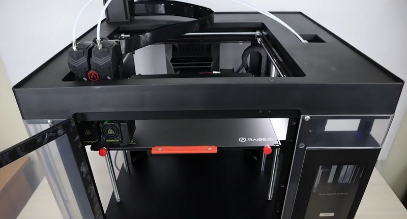 The Raise3D Pro3 HS 3D printer is shown from a top-down perspective, focusing on its internal structure. Visible details include a dual extruder system with filament feeding tubes, an open top section with clear panels, and a visible branding logo on the print bed. The image highlights the printer's robust frame, advanced filament handling, and accessibility features for ease of monitoring and maintenance.