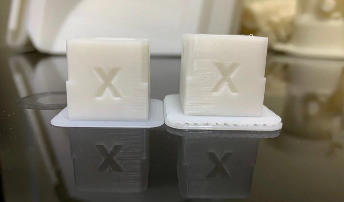 Two white 3D-printed cubes with raised "X" on surfaces, created by Raise3D Pro3 HS, reflect on a glossy black surface.