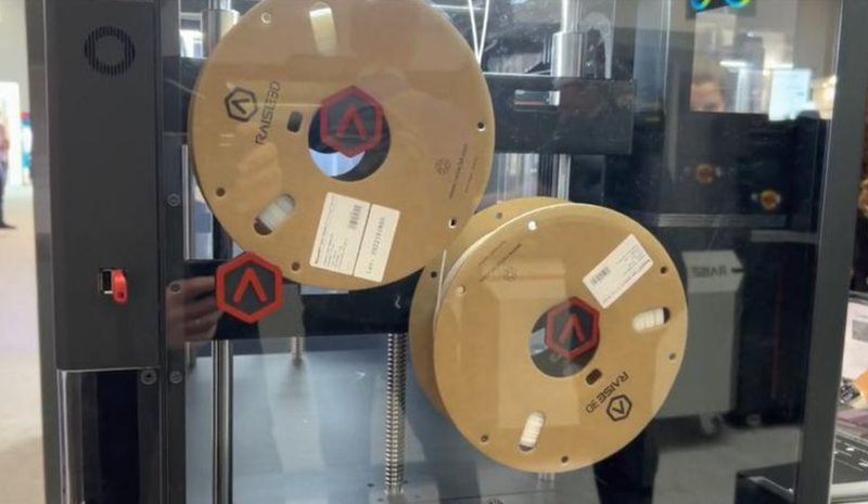 A close-up view of the Raise3D Pro3 HS 3D printer showcasing its filament feeding system. Two large spools of filament are mounted inside the transparent enclosure. The spools, labeled with the Raise3D branding, are positioned on a metal rod. The enclosure is sleek with a black frame and transparent panels, allowing a clear view of the internal components. A red switch is visible on the side, indicating user-friendly control access.