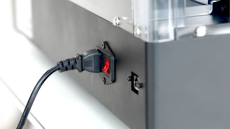 The image shows a close-up of the Raise3D Pro3 HS 3D printer's power connection. It highlights a black power cord plugged into a red power switch located on the printer's side panel. The panel is sleek and features a dark gray finish, complemented by clear plastic on the upper section, suggestive of a protective casing or enclosure part. The setting appears to be a modern and clean environment, emphasizing the printer's precision engineering and user-friendly design.