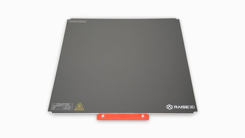The build plate from the Raise3D Pro3 HS, featuring a smooth, dark surface with a red handle at the bottom. Branding is visible in the lower right corner, with the Raise3D logo and safety warnings on the upper left. This component is essential for providing a stable, heated surface for successful 3D printing.