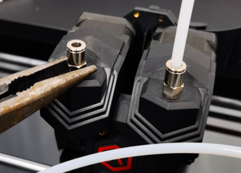 Close-up view of the Raise3D Pro3 HS 3D printer's dual extrusion heads. The image shows two robust, angular extruder covers with a metallic finish and geometric patterns. A pair of pliers is grasping one of the nozzles, likely for maintenance or replacement. A white filament is visible being fed into one of the nozzles, demonstrating the printer’s dual extrusion capability.