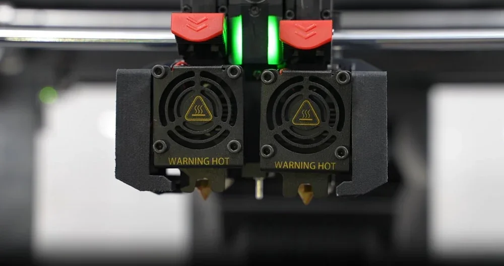 Raise3D Pro3 Plus dual extruder close-up with heat warning, black casing, red accents, and glowing green light, suggesting active filament printing.