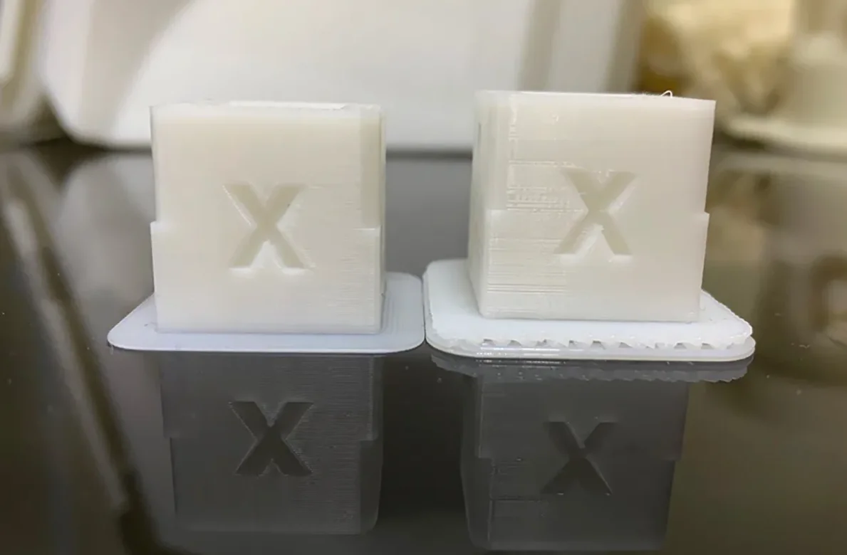 Two white 3D-printed cubes with embossed "X" on top, created by Raise3D Pro3 Plus, are set on a reflective surface for comparison.