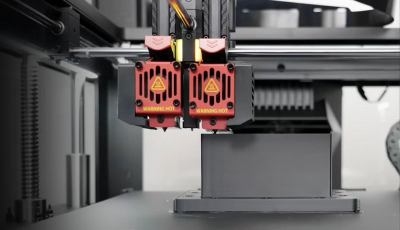 Close-up of the dual extruder system in the Raise3D Pro3 Plus HS 3D printer. The red extruders showcase clear "Warning Hot" labels, emphasizing the high-temperature operation. The black nozzles are positioned over a build platform, prepared for precise filament deposition. The image highlights the technical prowess and design of the printer, underscoring its capability for complex and efficient 3D printing tasks.