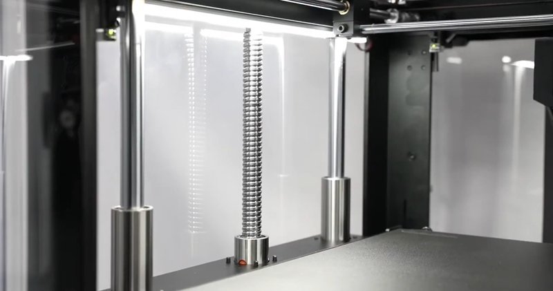 The internal view of the Raise3D Pro3 Plus HS 3D printer showcases the Z-axis mechanism, featuring a robust leadscrew surrounded by sleek, vertical guide rods. The reflective metal components and housing convey a durable and precise engineering design. Ambient lighting from above illuminates the setup, highlighting the smooth surfaces and intricate details of the printer assembly.