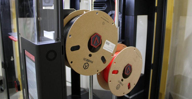 A close-up view of the filament spools used with the Raise3D Pro3 Plus HS 3D printer, situated adjacent to the printer’s touchscreen interface. The spools, branded with the Raise3D logo, are mounted on the printer’s side, one black and one orange, illustrating the dual-filament support. The setting highlights the printer's modern design and organized setup, integral for professional or industrial 3D printing environments.