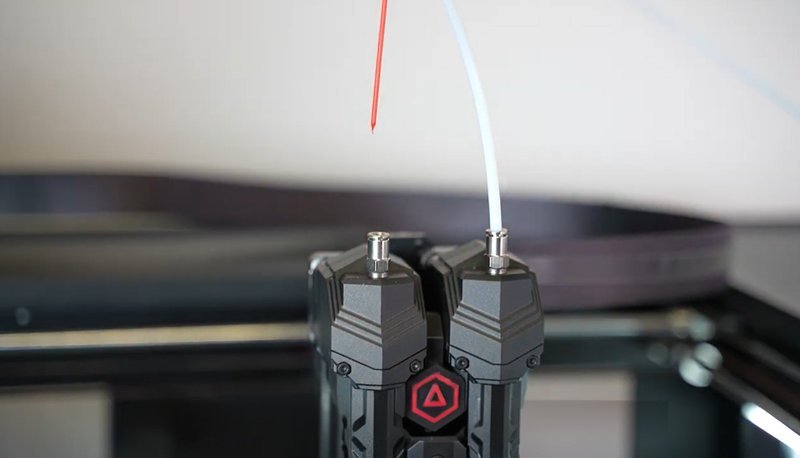 A close-up view of the Raise3D Pro3 Plus HS 3D printer’s dual extrusion system, showing the intricately designed nozzles. The nozzles are metallic with a detailed geometric pattern and are equipped with filament feed tubes—one red and one white—indicating multi-material or color printing capability. A red triangle-shaped logo is visible, emphasizing the brand's signature design on the extruder components.