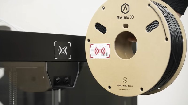 A close-up view of a filament spool used by the Raise3D Pro3 Plus HS 3D printer. The spool is branded with the Raise3D logo and features a built-in NFC tag, indicated by a wireless symbol on a sticker. The spool contains black filament, and the surrounding printer components are visible in a sleek, black finish, suggesting a modern design.